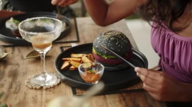 Restoranda hamburger ve patates kızartması yiyen zenci bir kadın. Bronzlaşmış kadın, otantik kafede akşam yemeği için patates dilimli siyah burger yiyor. Etnik kız iştah açıcı vejetaryen yemeğinin tadını çıkarıyor..