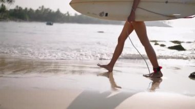 Okyanus sahilinde sörf tahtasıyla yürüyen Afrikalı Amerikalı kadın ayakları. Siyahi kadın sörfçü sörf tahtası kullanıyor. Çok ırklı bir kız gün doğumunda sörf seansına gidiyor. Yavaş çekim, düşük açı kenar görünümü.