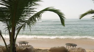 Ocean view with waves running, green palm trees and boho style cafe furniture in the foreground. Beautiful blue sea and calm waves float on the Indian Ocean. Background landscape
