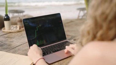 Dizüstü bilgisayardaki kadın kripto para tüccarı Candlestick çizelgelerini kontrol ediyor. Açık hava tropikal deniz kenarı kafesinde çalışıyor. Kadın kripto komisyoncusu hisse senedi kurlarını ve bahisleri okyanusa yakın hesaplıyor.