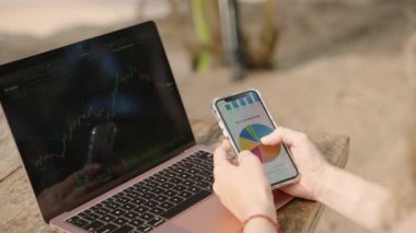 Akıllı telefondan istatistik çizelgelerini kontrol eden dizüstü bilgisayarı olan bir kadın kripto simsarı. Şifreli para birimleriyle çalışan serbest çalışan bir kadın telefonda diyagramları kontrol ediyor. Şifreleme grafiklerini yakından inceleyen kadın analist