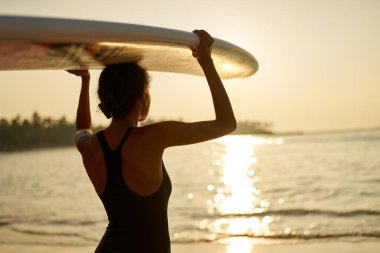 Okyanus sahilinde kafasında sörf tahtası olan Afrikalı Amerikalı bir kadın. Siyah sörf tahtası ile poz veren kadın sörfçü. Çok ırklı bir kız güneş doğarken sörf yapmaya gider. Güneşin tadını çıkarır..