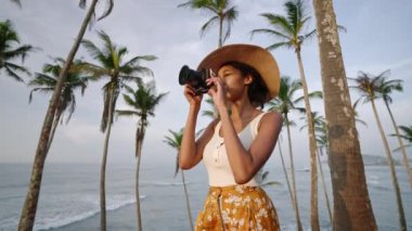 Afrikalı Amerikalı kadın tropikal adada kamerayla fotoğraf çekiyor. Kadın çok ırklı fotoğrafçı tatilde okyanus manzaralı yerleri keşfediyor. Siyah kız güneşin doğuşunda hasır şapkayla fotoğraf çekiyor.