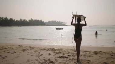 Kafasında sörf tahtasıyla okyanusa koşan Afrikalı Amerikalı sörfçü. Plajda sörf tahtası olan siyah bir kadın. Kız güneş doğarken tropikal bir yerde sörf yapmaya gidiyor. Yavaş çekim.