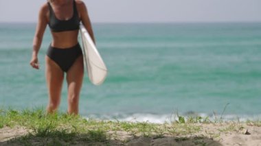 Güneşli bir günde, tropik plajda sörf tahtası taşıyan mutlu kadın sörfçü. Mayolu neşeli sörfçü kız su sporlarından zevk alıyor..