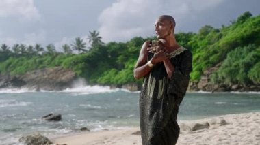 Çift cinsiyetli, lüks elbiseli eşcinsel bir adam okyanus sahilinde poz veriyor. Uzun gösterişli elbiseli ve aksesuarlı ikili olmayan model şık duruşlu kamera standlarına yavaşça bakıyor.