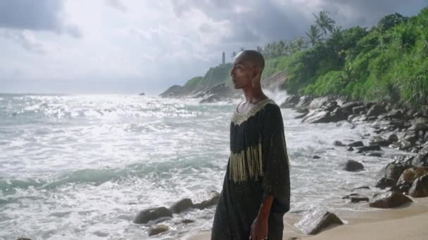 風光明媚な岩の海のビーチで豪華なドレスで男色の黒い人 トランス性的民族ファッションモデル身に着けているジュエリーでポッシュガウンを身に着けている劇的な空に対して熱帯の場所を歩く — ストック動画