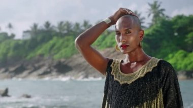 Lüks elbiseli gösterişli siyahi erkek, okyanus manzaralı bir sahilde poz veriyor. Uzun ve gösterişli bir elbise içinde transseksüel etnik model ve aksesuarlar kameraya bakar, boynuna yavaşça dokunur.