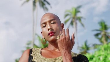 Androgynous abartılı eşcinsel model egzotik tropikal mekanlarda yakın çekim yapıyor. Cinsiyet akışkan etnik trans insan gökyüzüne karşı, palmiye ağaçları kollarını kavuşturmuş kameraya bakıyor.