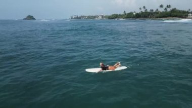 Genç bayan sörfçü güneşli bir günde deniz manzaralı tropik bir yerde sörf tahtasında kürek çekiyor. Sörfçü kız yaz tatilinde İHA 'dan sörf ve kürek çekmekten zevk alıyor..