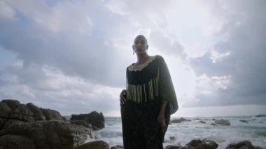 Renkli Androgynous kameraya bakıyor. Lüks elbiseli, kayalık tropikal okyanus sahilinde takılar takıyor. Transseksüel manken burun halkası piercing, yüzük, küpe takıyor..