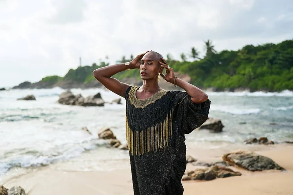 Epatage lgbtq black male posing with hands up on camera on scenic ocean beach. Non-binary ethnic fashion model in long posh dress wears jewellery stands gracefully on sea shore and a lighthouse.