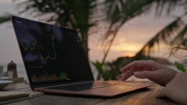 Woman crypto trader at laptop checking candlestick charts online working remotely at outdoor tropical seaside cafe. Female broker analyses cryptocurrency exchange rates on crypto stock by ocean