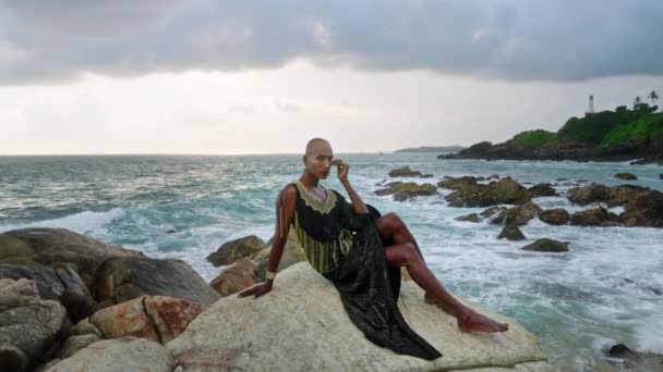 豪華なドレスでゲイ二人組の男 ブティックの真鍮の宝石類 化粧ポーズは熱帯岩の海のビーチに座っている ポッシュガウン 鼻リングピアス イヤリング 宝石でリングで同性愛のページファッションモデル — ストック動画