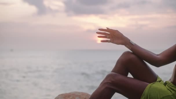 オープンドレスのLgbtqia黒人は ジュエリーは岩の上に座って 美しい海の海岸線の夕日に手で太陽に触れます ジェンダー流体民族神モデルは 黄金のアクセサリーを身に着けている 宝石リング ブレスレット — ストック動画