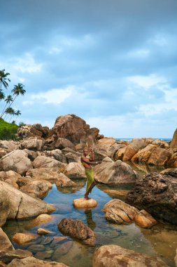 İkili olmayan siyah kişi bir derenin içindeki durgun suyun ortasında bir kayanın üzerinde duruyor. Lgbtq etnik ve zarif model, tropikal bölgede kristal berrak bir gölette bir taşın üzerinde duruyor..