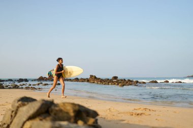 Okyanus sahilinde sörf tahtasıyla koşan Afrikalı Amerikalı bir kadın. Sörf tahtası olan siyah bir kadın sörfçü. Çok ırklı bir kız güneş doğarken tropikal bir yerde sörf yapmaya gidiyor..
