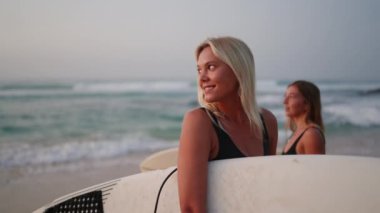 Gün doğumunda sahilde sörf tahtası tutan gülümseyen sörfçü kızlar. Neşeli dişi sörfçüler şafakta okyanusun kenarında dururlar. Deniz kenarında sörf yapan genç kadınlar ve dalgalar, konuşmak, gülmek, yer aramak.