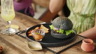 Önünde hamburger ve patates kızartması olan bir kadın. Bir açık hava restoranının masasında patates dilimleri ile servis edilen siyah burger. Otantik kafede akşam yemeği için iştah açıcı vegan yemeği..