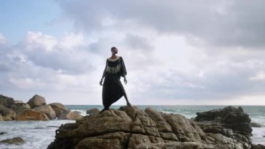 Renkli Androgynous, okyanus kıyısındaki kayalıklarda butik şık elbiseler giyiyor. Efsanevi kıyafetli transseksüel etnik model altın mücevher takıyor, Oshun tanrıçası konsepti gibi poz veriyor..