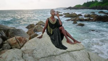 Ahlaksız LGBTQ bipoc çalışanı lüks elbiseler, butik pirinç mücevherler içinde dalgalı okyanuslara karşı uçurumda poz veriyor. Sosyetik cüppeli çok ırklı bir model. Burun halkası piercing, küpe, yüzük takıyor..