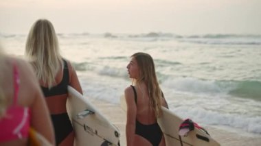 Gün doğumunda sahilde yürüyen sörfçü kızlar. Sörf tahtaları taşıyan dişi sörfçüler şafak vakti okyanus boyunca sahilde yürüyorlar. Çok ırklı genç sörfçü kadınlar okyanus akıntısında geriye doğru yürüyorlar.