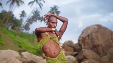 Açık elbiseli, altın küpeli, burunlu, bilezik takan, avuç içlerine, uçurumlara, tropiklere karşı elleri havada poz veren ikiz siyahi olmayan biri. Queer lgbtq Moda Modeli kokteyl kıyafeti giyer..