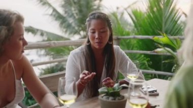 Çeşitli bayan arkadaşlar gün batımında deniz manzaralı bir restoranda şarap içerler. Çok ırklı kadınlar konuşur, alacakaranlıkta tropikal bir kafede gülümserler. Çok ırklı genç kadınlar yemek yiyor. Palmiye ağaçları, arka planda okyanus.