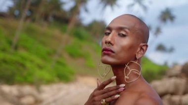 Renkli Androgynous, kayalık okyanus kıyısındaki kayalıklar arasında açık bir elbise giyer. Dar elbiseli transseksüel etnik model el yapımı mücevherler, burun halkası, küpeler, yüzükler takıyor.