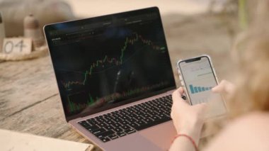 Akıllı telefondan istatistik çizelgelerini kontrol eden bilgisayardaki kadın kripto tüccarı. Şifreli para birimiyle uzaktan çalışan bir kadın telefonda şema kontrol ediyor. Şifreleme grafiklerini yakından inceleyen kadın analist