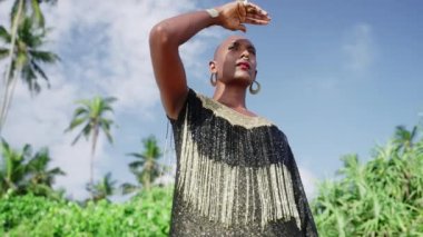 Butik elbise, altın küpeler, burun halkası, bilezik, bilezik, güneş ışığıyla kaplı elleri tropikal palmiye ağacı plajında. Queer lgbtq modeli ışıltılı elbiseyle çok uzakta görünüyor..