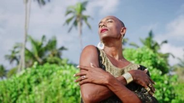Lüks elbiseli, kadın makyajlı Androgynous, tropiklerde poz veriyor. Parlak elbisesi, mücevherleri ve palmiye ağaçlarına karşı elleri ile kendini kucaklayan transseksüel etnik model..