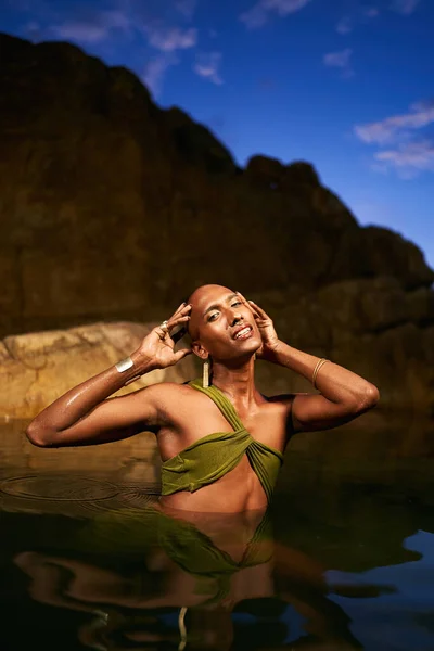 stock image Happy trans sexual fashion model shows golden jewelry chest deep inside natural pool surrouded by rocks at night. Androgynous ethnic person stands in the middle of backwater exotic scenic location