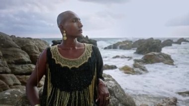 Lüks elbiseli eşcinsel bipoc adam, butik pirinç mücevher, tropik kayalık okyanus sahilinde otururken makyaj pozları. Homoseksüel epatage mankeni, gösterişli bir elbise, piercing, küpeler, mücevherli yüzükler..