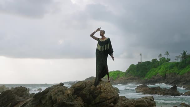 海の海岸線の上の岩のブティックのポッシュドレスには 色の異様な人が立っています 壮大な衣装でトランス性的民族ファッションモデルは黄金のジュエリーを身に着けています Oshun女神の概念のようなポーズ — ストック動画