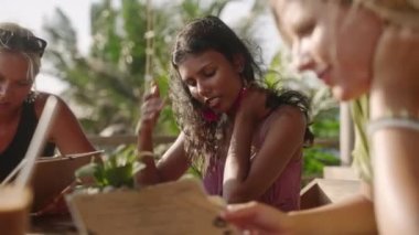 Açık hava lokantası menüsündeki çeşitli kadın arkadaşlar. Tropikal bir kafede oturan çok ırklı kızlar birbirleriyle konuşarak yemek seçiyorlar. Genç çoklu etnik kadınlar yemek yiyor.