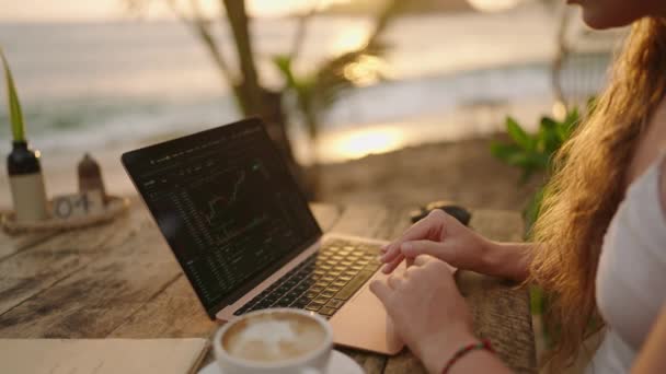 ラップトップの女性暗号通貨トレーダーは 日没時に屋外の熱帯海辺のカフェでリモートで働いているチャートをオンラインでチェックします 女性暗号ブローカーは 海のクローズアップによる株式為替レートのグラフィックを分析します — ストック動画
