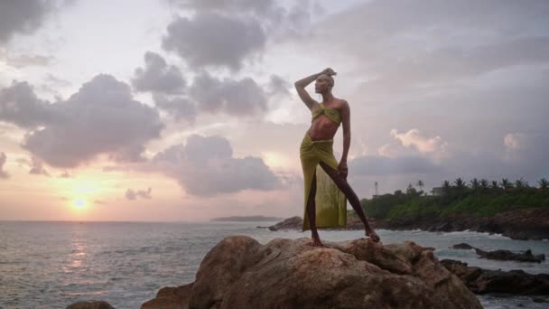 ブティックドレスの神の非バイナリ黒人 劇的な海の夕日のスカイラインの上の岩の丘の上に真鍮の宝石類のポーズ 空気になびくオープン衣装でクィアLgbtqファッションモデル パンテオンの概念 — ストック動画