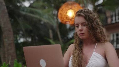 Focused thoughtful woman at outdoor tropical location looking at laptop learning, working on project online. Concentrated serious female thinking of problem solution, works remotely on computer
