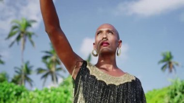 Lüks elbiseli eşcinsel bipoc adam, butik pirinç mücevher, palmiye gölgesiyle kaplanmış makyaj örtüsü. Homoseksüel, etnik, şık bir cübbe giyen, burun halkası takan, piercing takan, küpe takan, gözlerini kaçıran biri. Palmiye ağaçları.