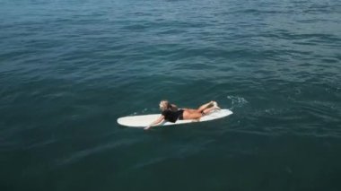 Genç bayan sörfçü güneşli bir günde deniz manzaralı tropik bir yerde sörf tahtasında kürek çekiyor. Sörfçü kız yaz tatilinde İHA 'dan sörf ve kürek çekmekten zevk alıyor..