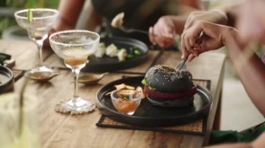 Restoranda hamburger ve patates kızartması yiyen zenci bir kadın. Bronzlaşmış kadın siyah burger yiyor ve patates dilimleri ile otantik kafede çatal ve bıçakla yemek yiyor. Etnik kız vejetaryen yemeğinden hoşlanıyor.
