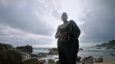 Lüks elbiseli gösterişli siyahi erkek, kayalık, fırtınalı okyanus sahillerinde poz veriyor. Uzun sosyetik elbiseli ve aksesuarlı transseksüel etnik model kameraya bakıyor, yavaş çekimde dramatik bir siluet.