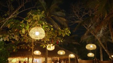 Geceleri ağaçlardan sarkan hasır lambaları tropikal arka planda süsleme olarak kullanılıyor. Dışarıdaki kafede eski moda ahşap ampuller sallanıyor. Egzotik ortamda Boho düğün süslemeleri.