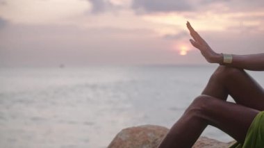 İlahi olmayan siyah insan eli, üzerinde pirinç mücevherlerle güneşe karşı hareket eder dramatik okyanus günbatımı ufkunun üzerindeki kayalık tepede poz verir. Açık kıyafetli Queer lgbtq model bir tepede oturuyor..