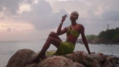 Lüks elbiseli eşcinsel bipoc adam, butik pirinç mücevher, tropik kayalık okyanus sahilinde otururken makyaj pozları. Homoseksüel epatage mankeni, gösterişli bir elbise, piercing, küpeler, mücevherli yüzükler..