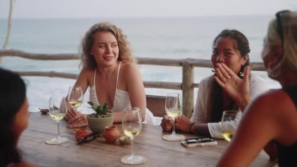 多人種の女性の友人は日没時に屋上のシービューレストランでワインを飲む 多様な女性が話し 夕暮れ時のトロピカルカフェで笑顔 若い多民族女性の食事 ヤシの木 背景に海 — ストック動画