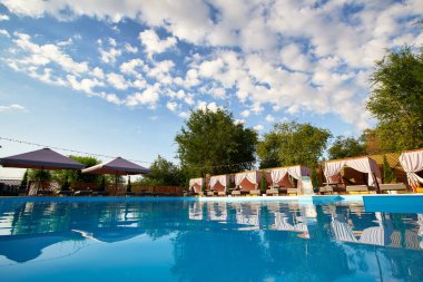 Etrafı yeşillik ve mavi suyla çevrili yazlık evler. Havuz kenarında çardaklar, şezlonglar ve güneş şemsiyeleriyle dolu havuz alanı. Tatilde huzur ve rahatlama ortamı.