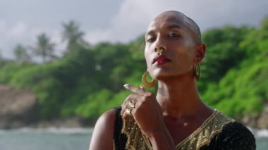 Butik elbise giymiş, altın küpeler takmış, deniz manzaralı sahilde ağır çekimde poz veren ikili olmayan siyahi biri. Queer lgbtq patronluk taslayan manken takı yüzükleri, tropiklerde gösterişli bir elbise giymiş..