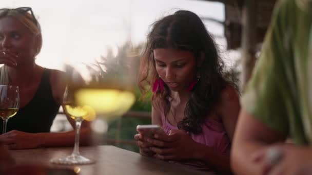 カラーの女性は スマートフォンのスクロールと入力を保持 友人と座って屋外の熱帯屋上カフェで彼女の携帯電話を使用してインドの女性 インターネットと携帯電話中毒 オンラインコミュニケーション — ストック動画